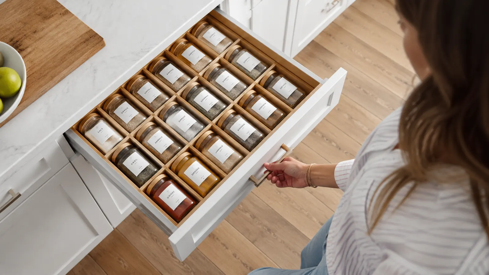 Woman opening kitchen drawer to reveal a neatly arranged in-drawer spice insert with labeled jar lids visible from above