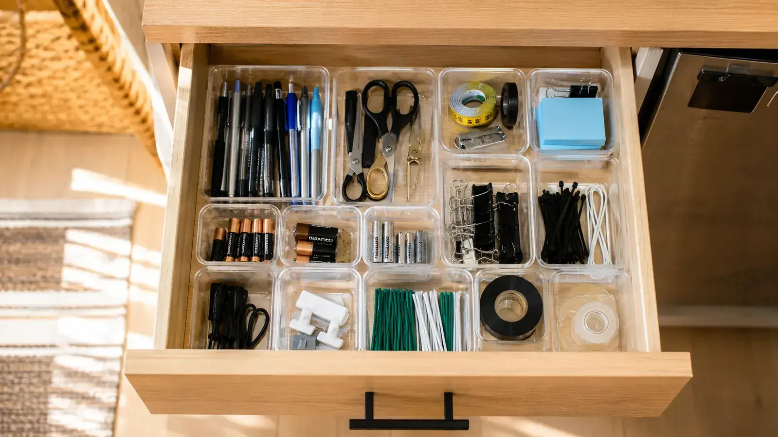 An organized kitchen junk drawer with clear plastic divider bins holding batteries, pens, tape, and small tools in neat labeled rows