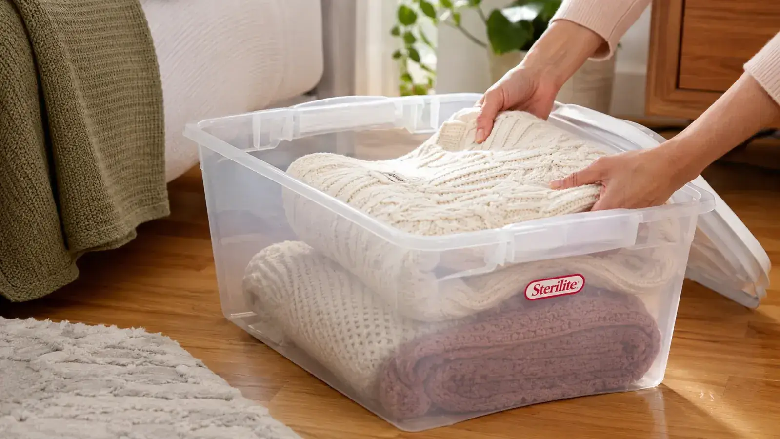 Hands placing a folded sweater into an open clear Sterilite latching bin on a bedroom floor
